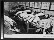 Men are depicted lying in a row on a long table in a room. One man is shown with a pipe in his mouth.