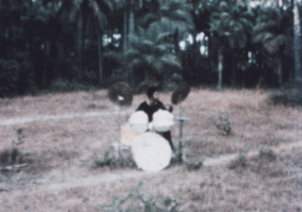 Tony Williams plays a drum kit in Senegal in a field with palm trees behind him